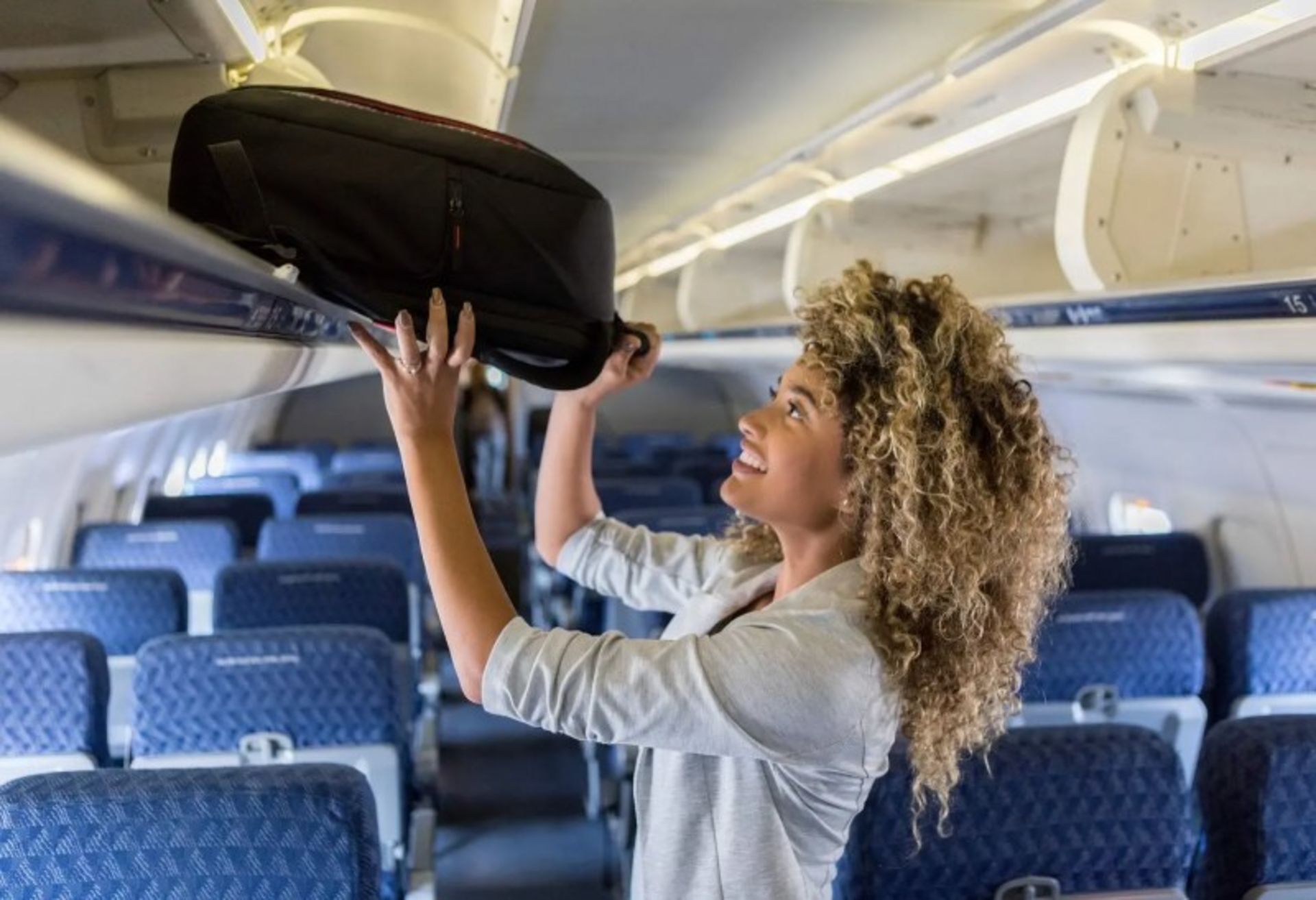 A woman with a curly hair is putting a black suitcase in the wardrobe over the plane