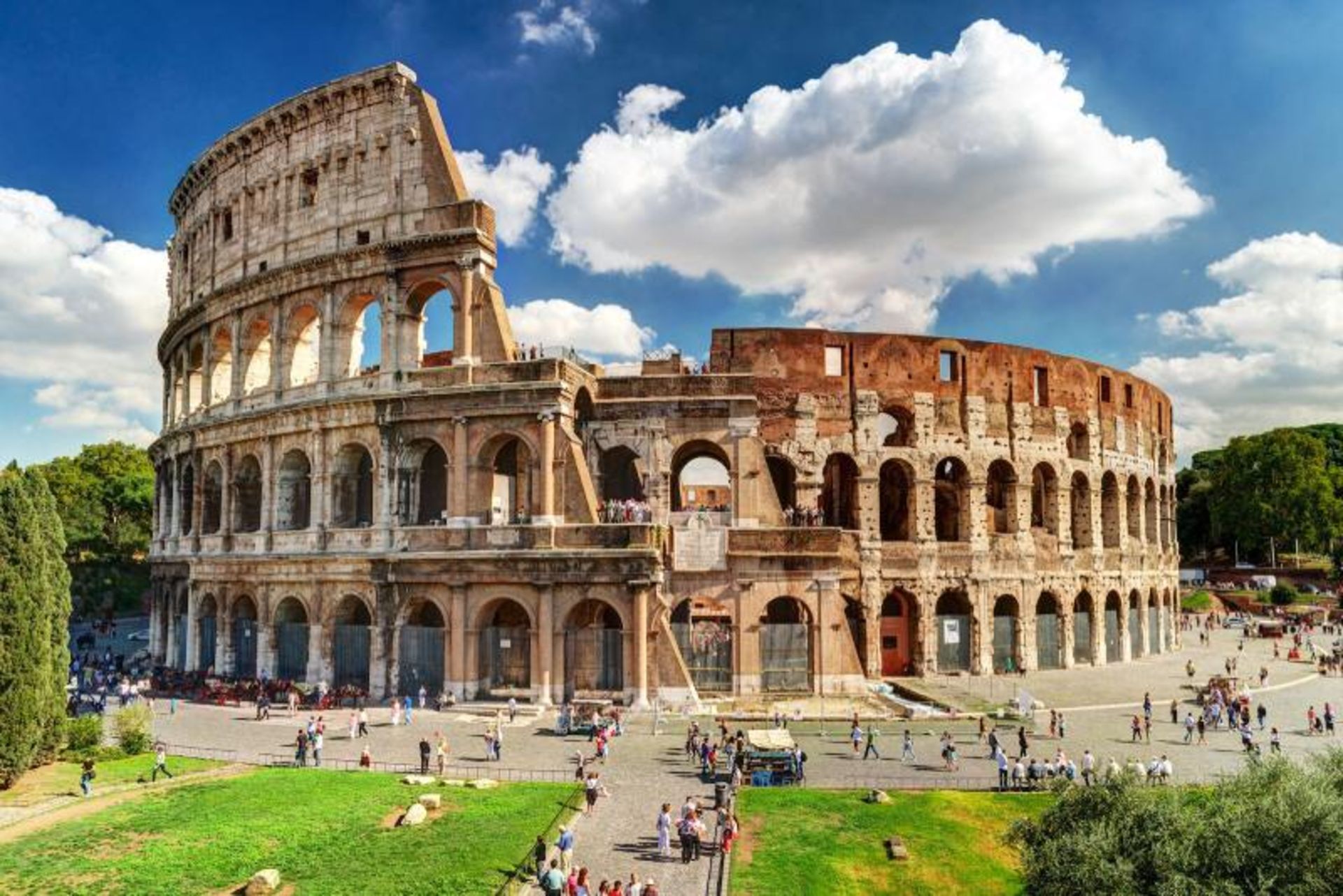 Colosseum amphitheater on sunny day, blue and cloudy sky, surrounding green space and visitors