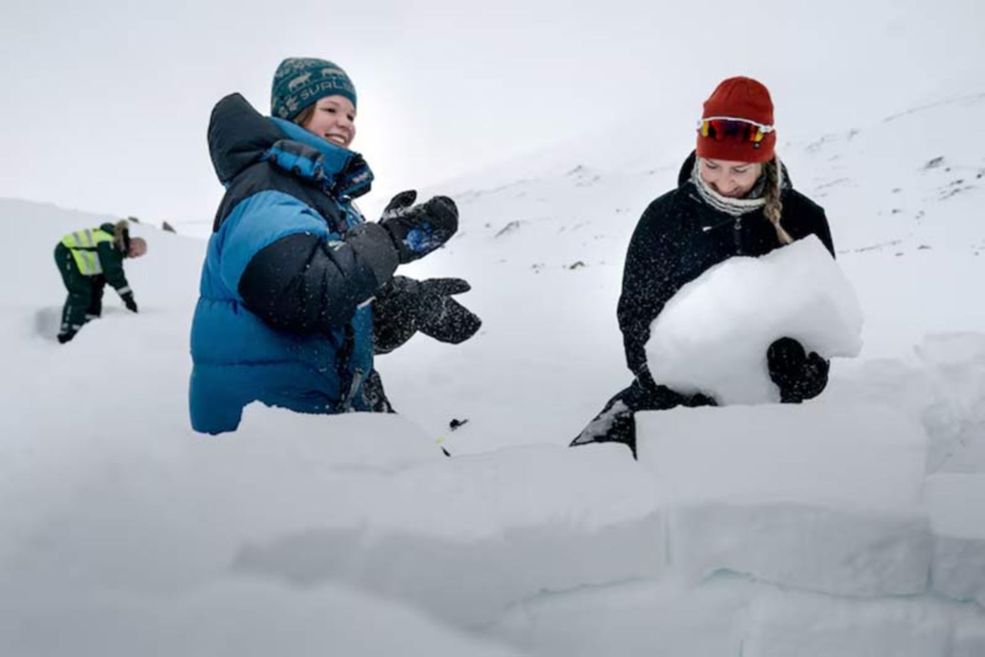 Two people in warm winter clothes building an ice house in the snow