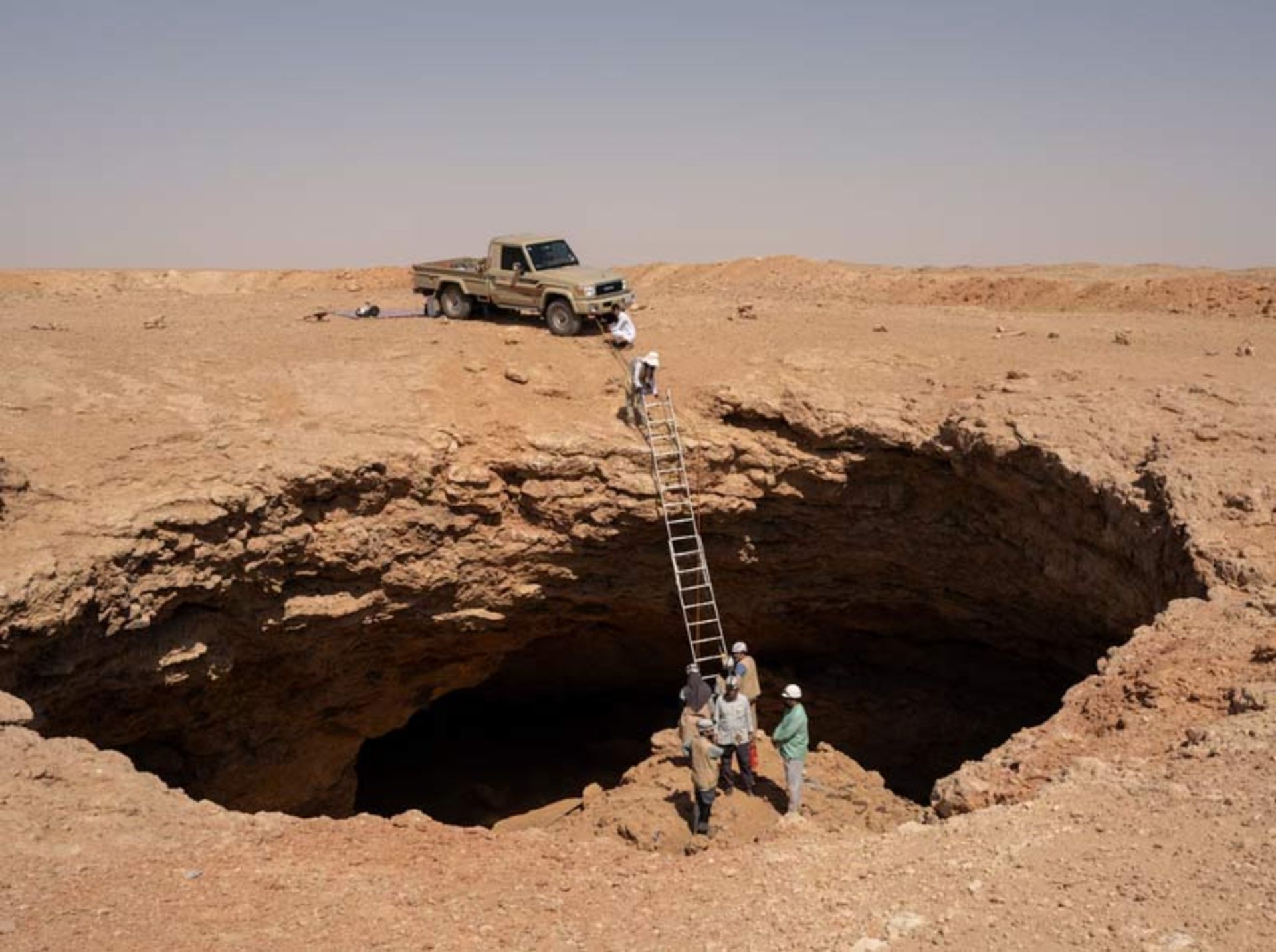 A car at the edge of a desert pit and a ladder leading into its dark depths